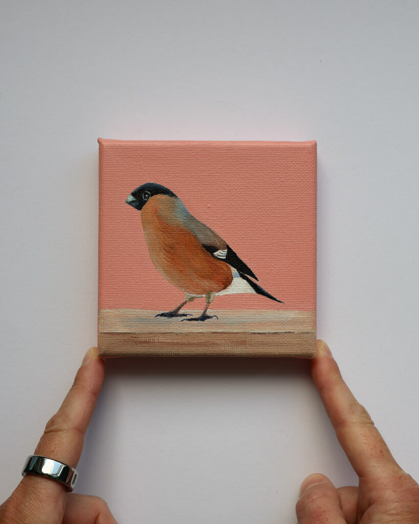 Klein schilderij van een vrouwtjesgoudvink op een houten rand tegen een warme koraalroze achtergrond